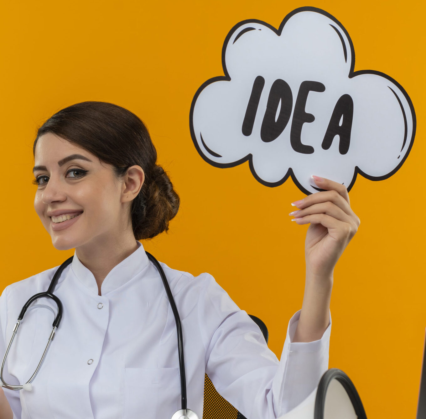 smiling young female doctor wearing medical robe and stethoscope sitting at desk with medical tools speaker and laptop holding idea bubble showing empty hand isolated on yellow background